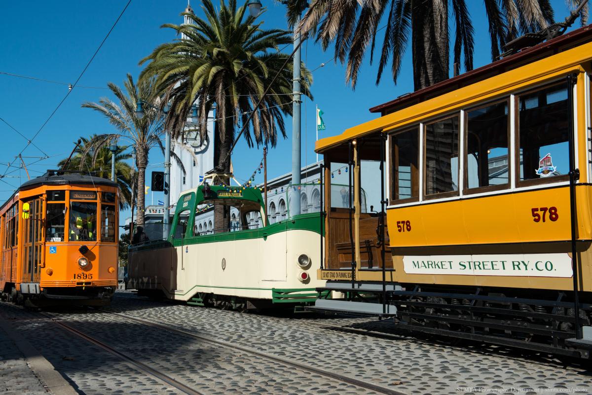 An orange historic streetcar passes a green and cream open-air Boat Tram and another orange historic streetcar. Palm trees and Ferry Building in the background. An orange historic streetcar passes a green and cream open-air Boat Tram and another orange historic streetcar. Palm trees and Ferry Building in the background.
