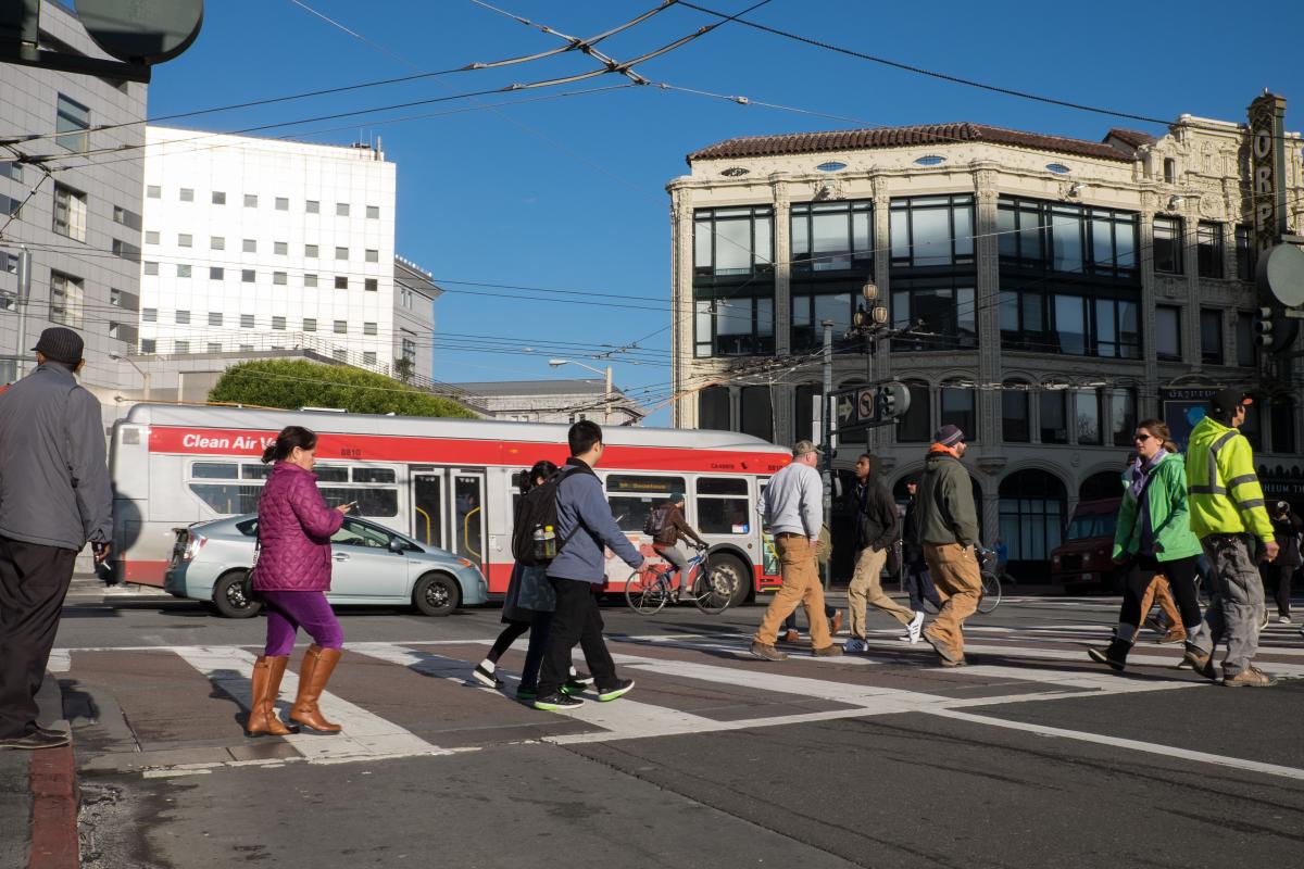People cross a street as cars and buses and people riding bikes travel in the lanes near them. People cross a street as cars and buses and people riding bikes travel in the lanes near them.