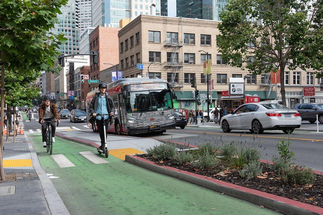 Scooter riders use a bikeway that's painted green on a city street. Buses and cars are in the background. Scooter riders use a bikeway that's painted green on a city street. Buses and cars are in the background.