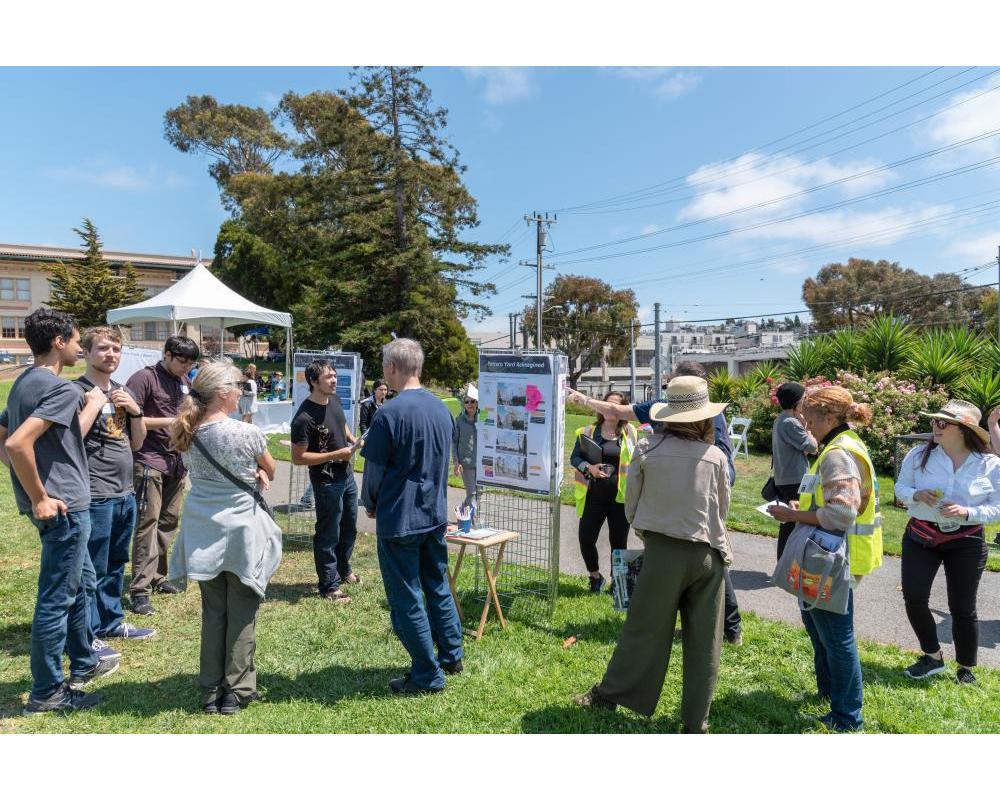 Image shows a group of people standing on a grassy hill, chatting near poster boards about Potrero Yard. Image shows a group of people standing on a grassy hill, chatting near poster boards about Potrero Yard.