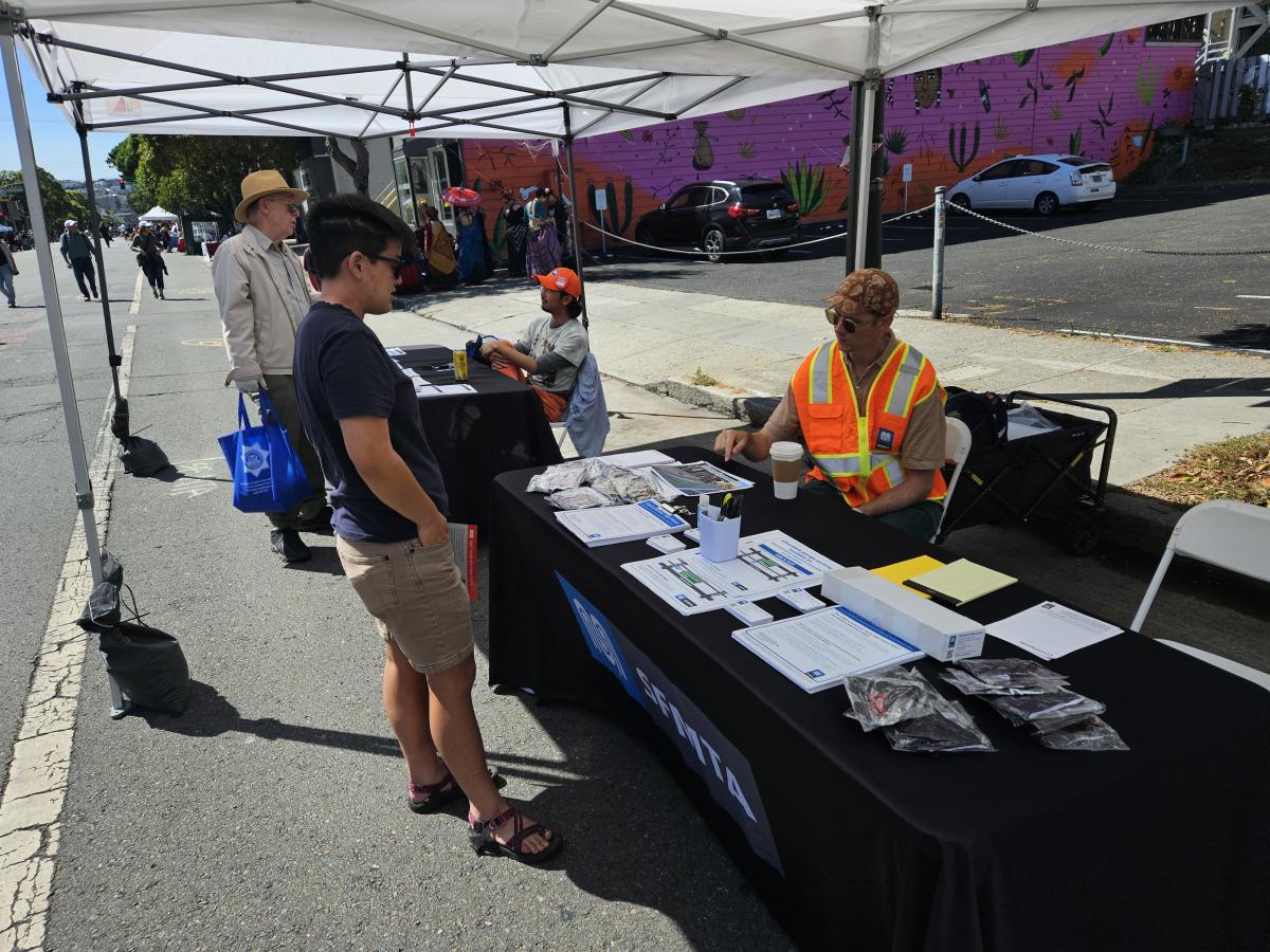 A member of the public is shown talking to an SFMTA staff person wearing an orange safety vest. A table, between them, is covered with diagrams and papers about the project. A member of the public is shown talking to an SFMTA staff person wearing an orange safety vest. A table, between them, is covered with diagrams and papers about the project.