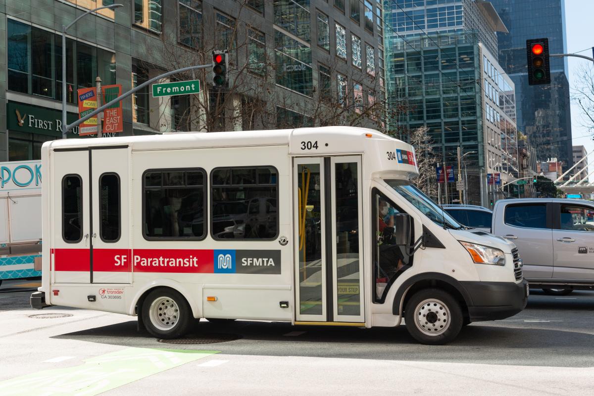 A SF Paratransit van crosses a downtown intersection along with other vehicles. A SF Paratransit van crosses a downtown intersection along with other vehicles.