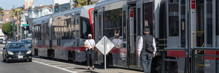 Riders board the K Ingleside train. The boarding island is too short, so one rider walks in the street next to the train.