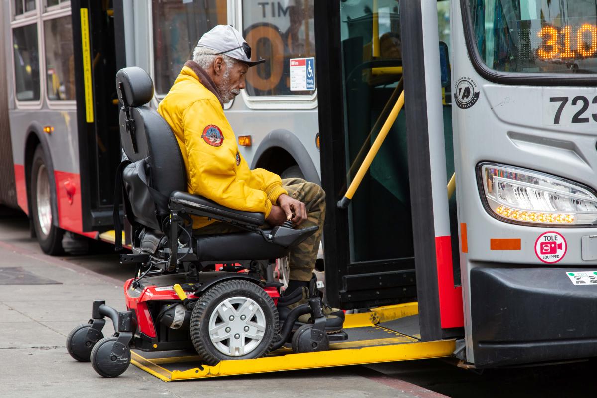 Person using a mobility device boards a Muni bus through a ramp lowered to sidewalk level. Person using a mobility device boards a Muni bus through a ramp lowered to sidewalk level.
