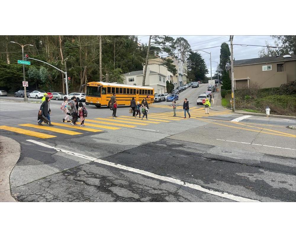 students using a crosswalk to cross clarendon avenue at panorama drive a bus is waiting in the intersection.
