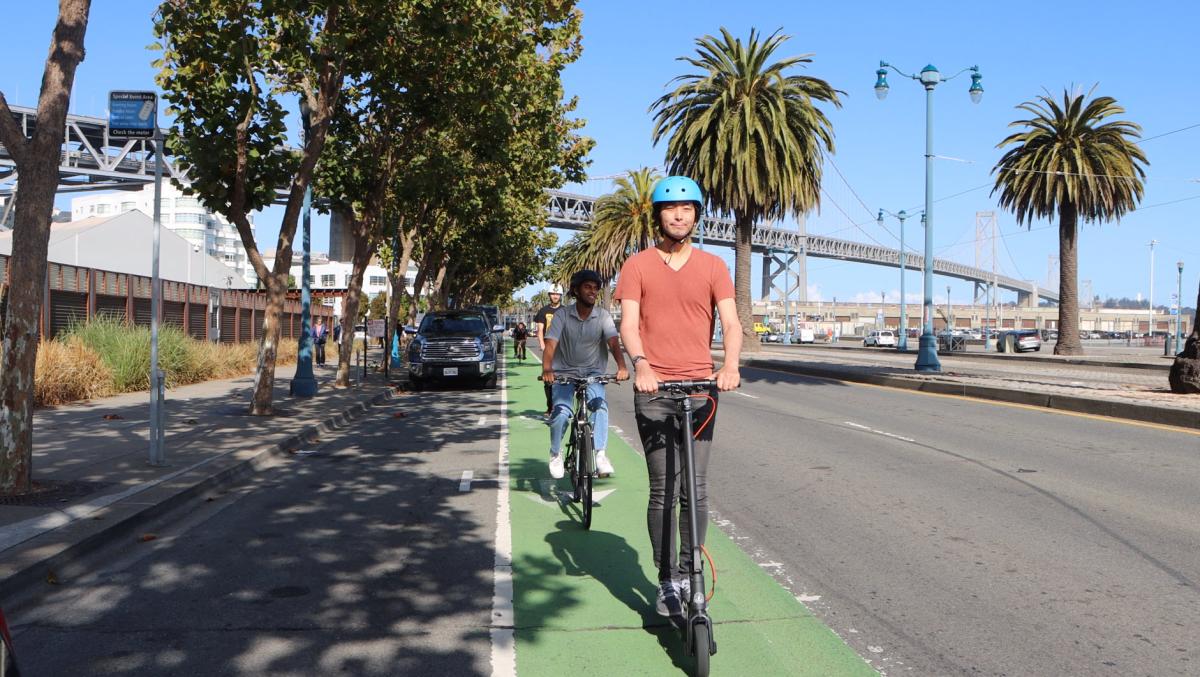 Two people ride scooters in a bikeway that's painted green by the Embarcadero. Two people ride scooters in a bikeway that's painted green by the Embarcadero.